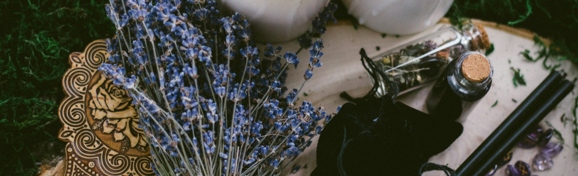 Des fleurs séchées, deux pots blanc, des fioles de verre contenant des herbes séchées, un symbole sur du bois gravé, le tout posé sur de l'herbe.